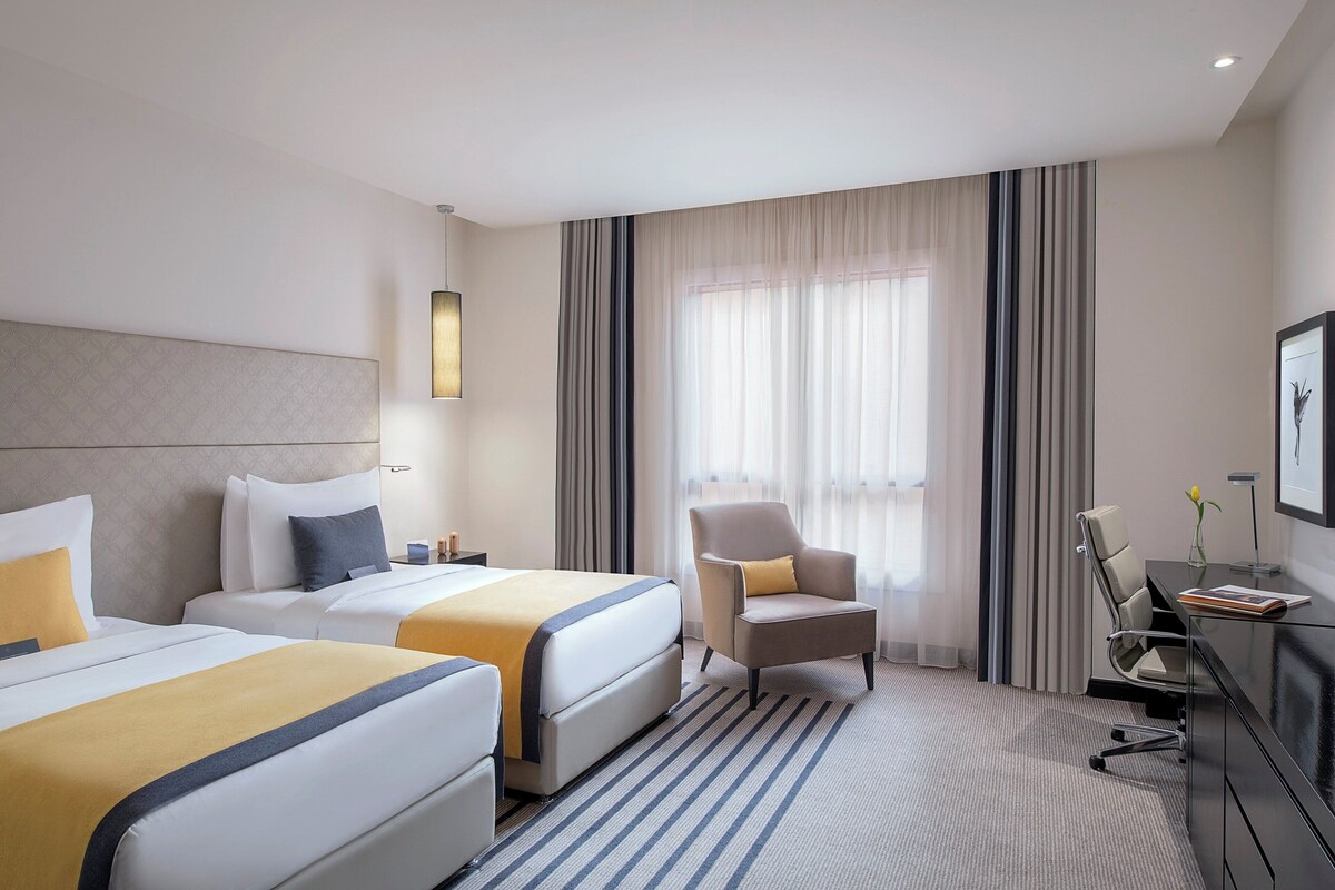 A hotel room with two beds, a desk, a chair, and a large window with curtains.