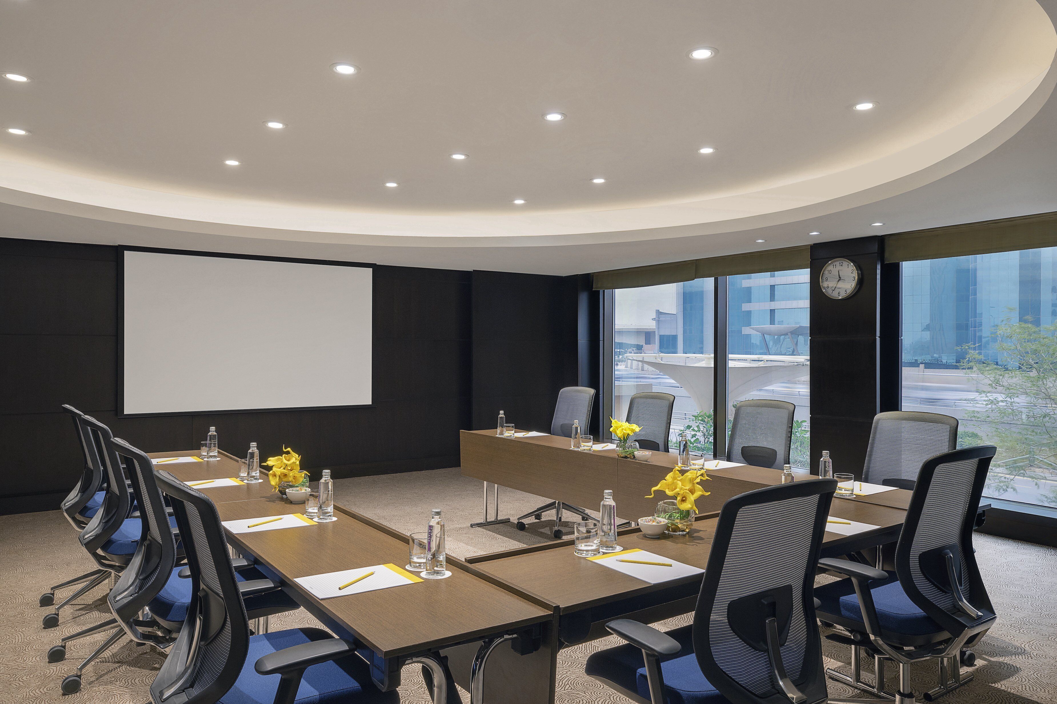 A conference room with a long table, chairs, a projector screen, and a clock on the wall.