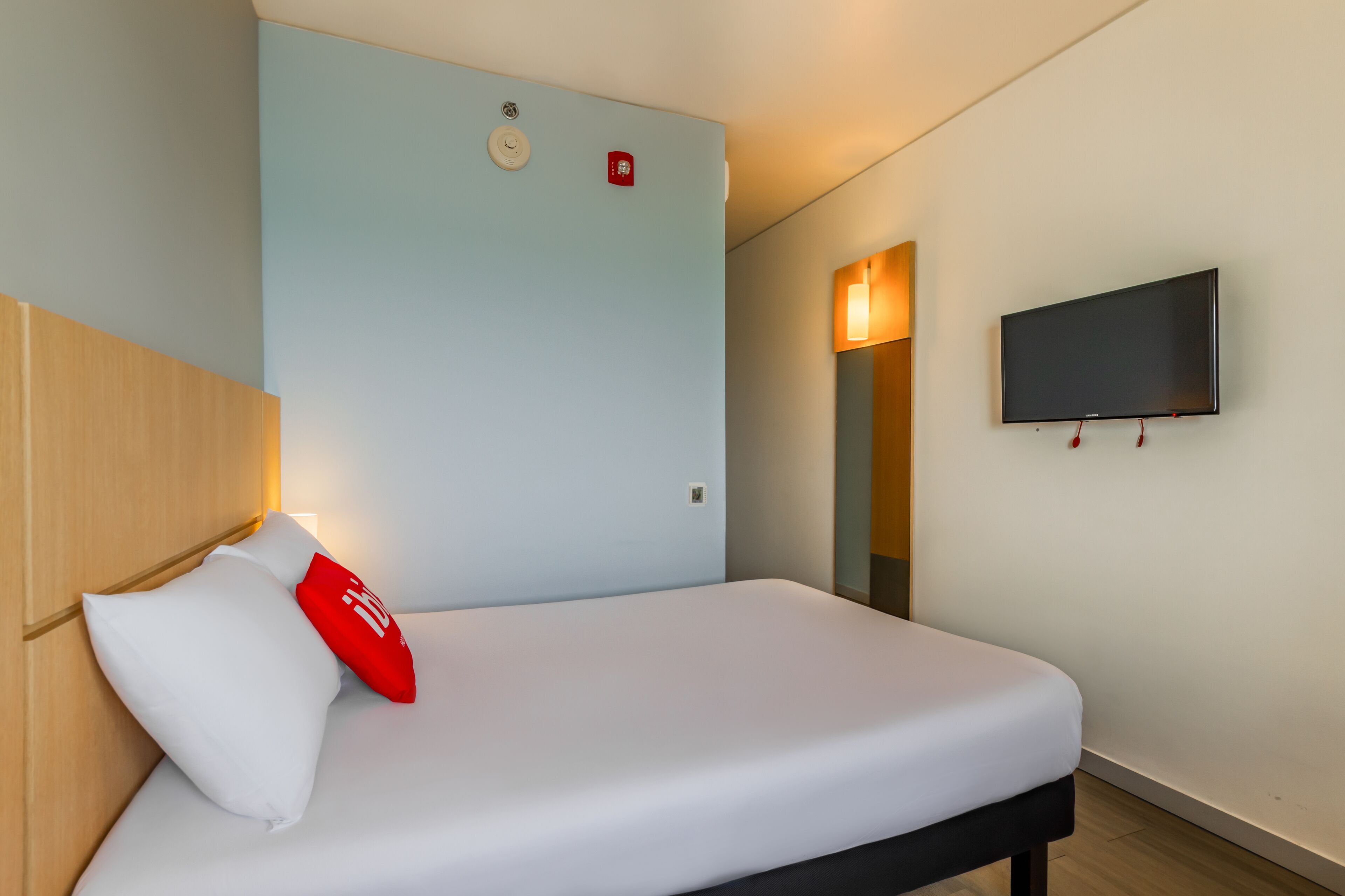 Una habitación de hotel con una cama, un televisor montado y una lámpara fijada a la pared.