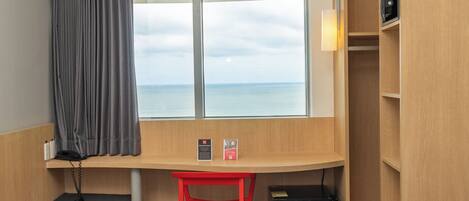 Ein Hotelzimmer mit Bett, Schreibtisch, Stuhl, Fenster mit Meerblick und einer kleinen Mülltonne.