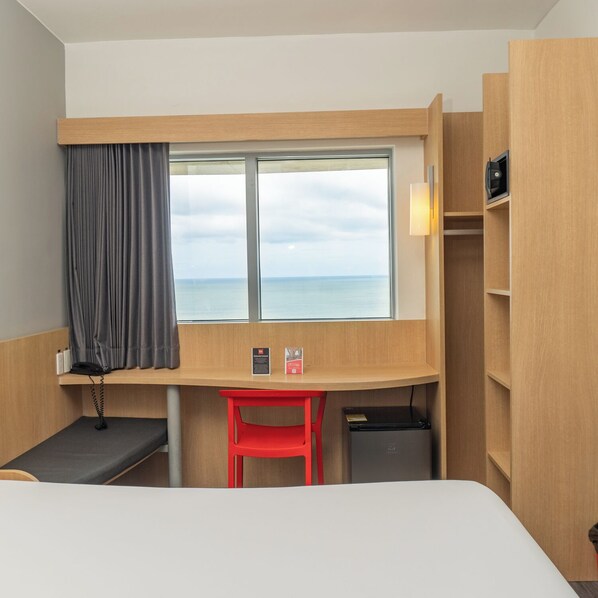 A hotel room with a bed, a desk, a chair, a window with a view of the sea, and a small trash bin. - ibis Cartagena Marbella (Cartagena)