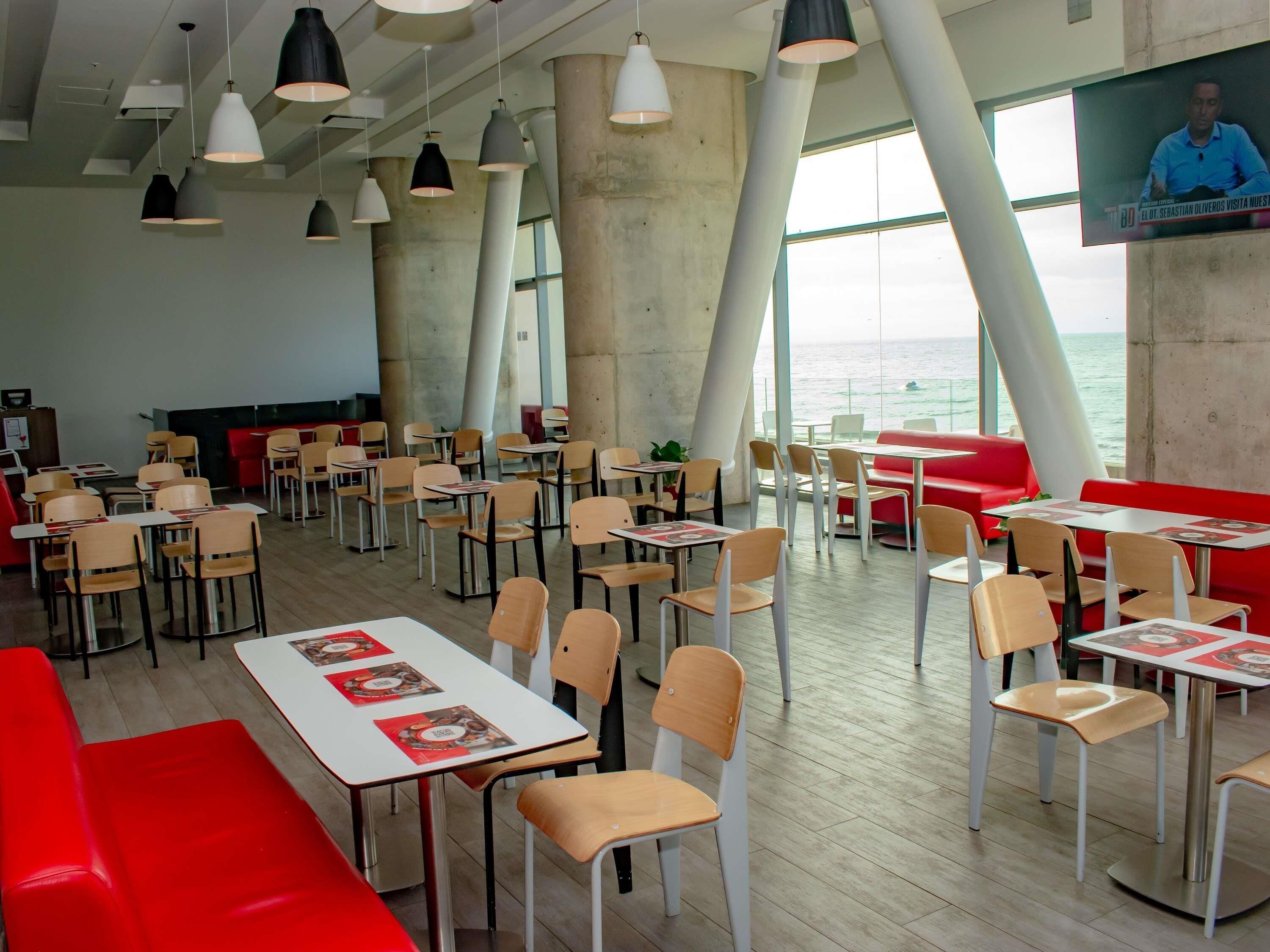 A modern dining area with tables and chairs, large windows, and a view of the sea.