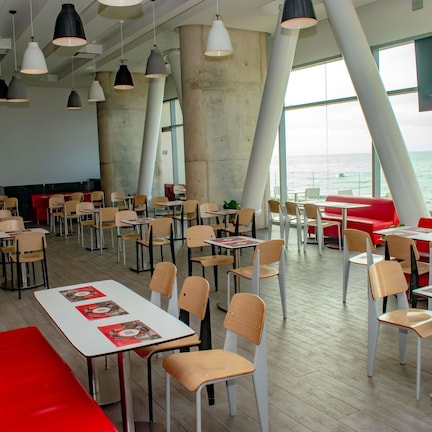 A modern dining area with tables and chairs, large windows, and a view of the sea.