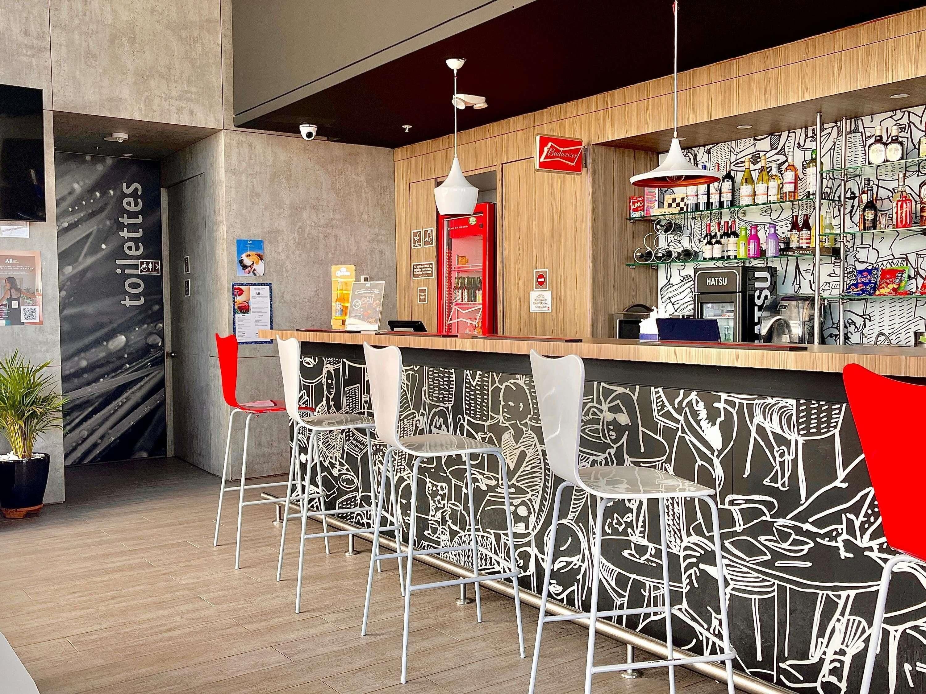 Una zona de bar con mostrador de madera, taburetes blancos y una pared con mural blanco y negro.