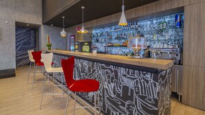 Un espace bar moderne avec un comptoir, des chaises rouges et un mur orné d’une fresque représentant des personnes en train de boire.