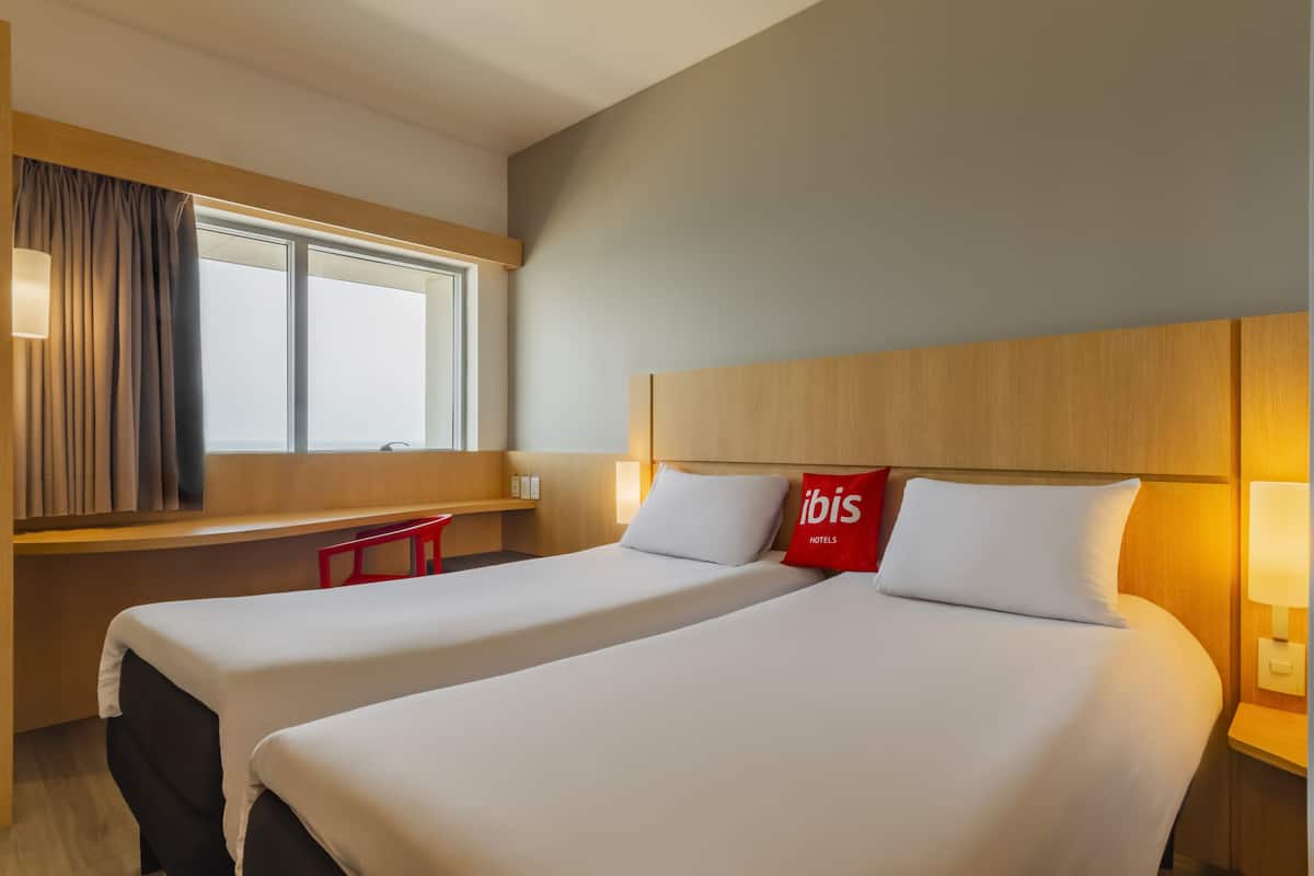 Et hotelværelse med to senge, et vindue med gardiner, en træsenggavl og en rød pude med 'ibis'-logo.