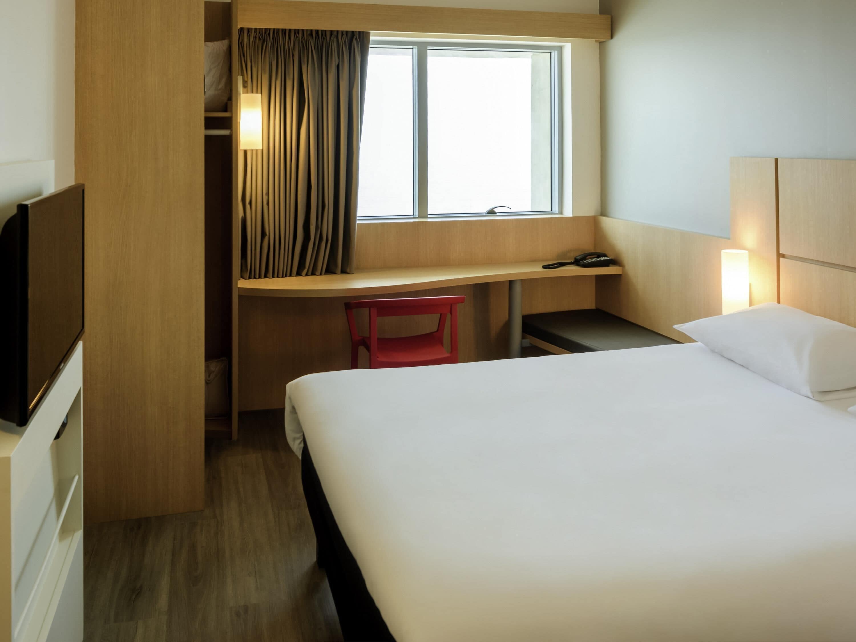 Habitación de hotel con cama, escritorio, silla, televisión, ventana con cortinas y lámpara de noche.