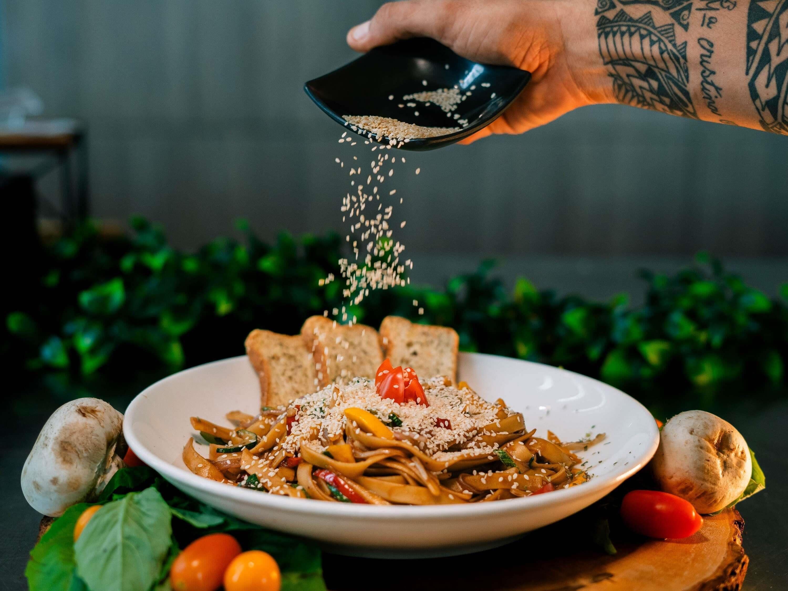 A plate of noodles garnished with sesame seeds and a cherry tomato, with a hand sprinkling more seeds.