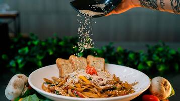 A plate of noodles garnished with sesame seeds and a cherry tomato, with a hand sprinkling more seeds.