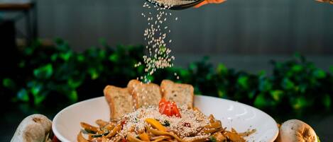 A plate of noodles garnished with sesame seeds and a cherry tomato, with a hand sprinkling more seeds.