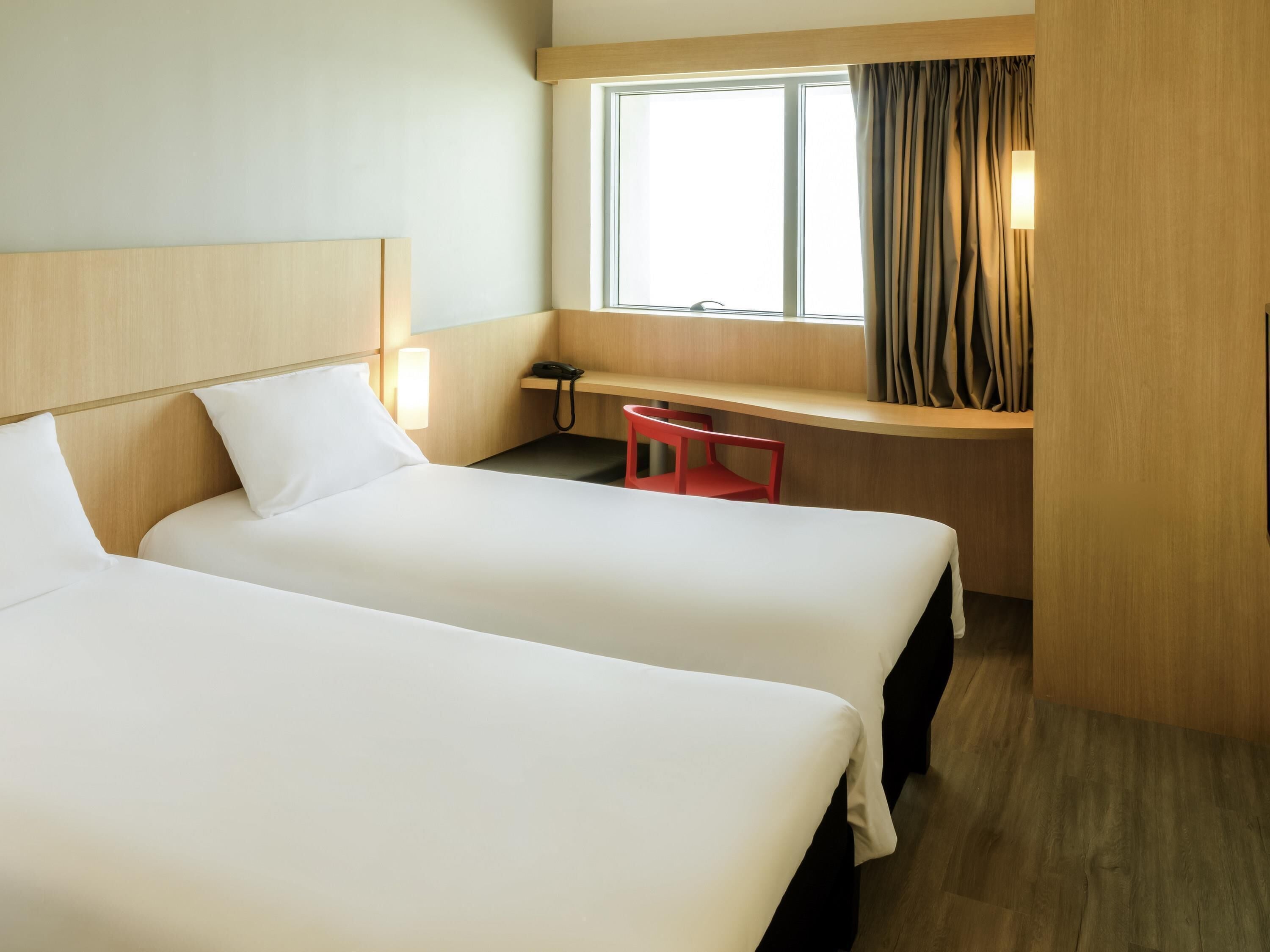 Habitación de hotel con dos camas, un escritorio, una silla, una ventana con cortinas y una lámpara de noche.