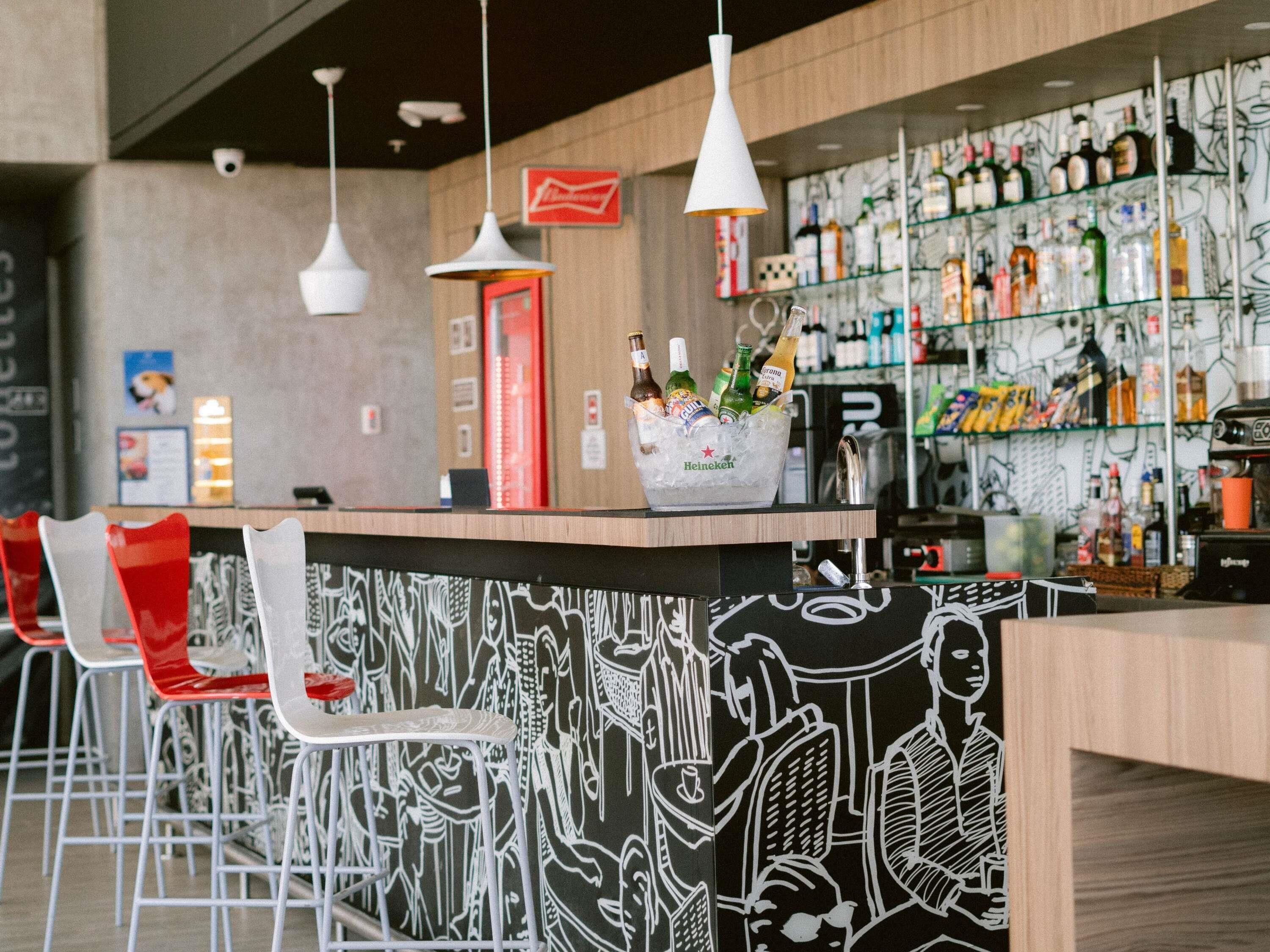Uma área de bar com balcão, balde de gelo e diversos tipos de garrafas. Há cadeiras vermelhas e brancas, e uma parede com um mural em preto e branco.