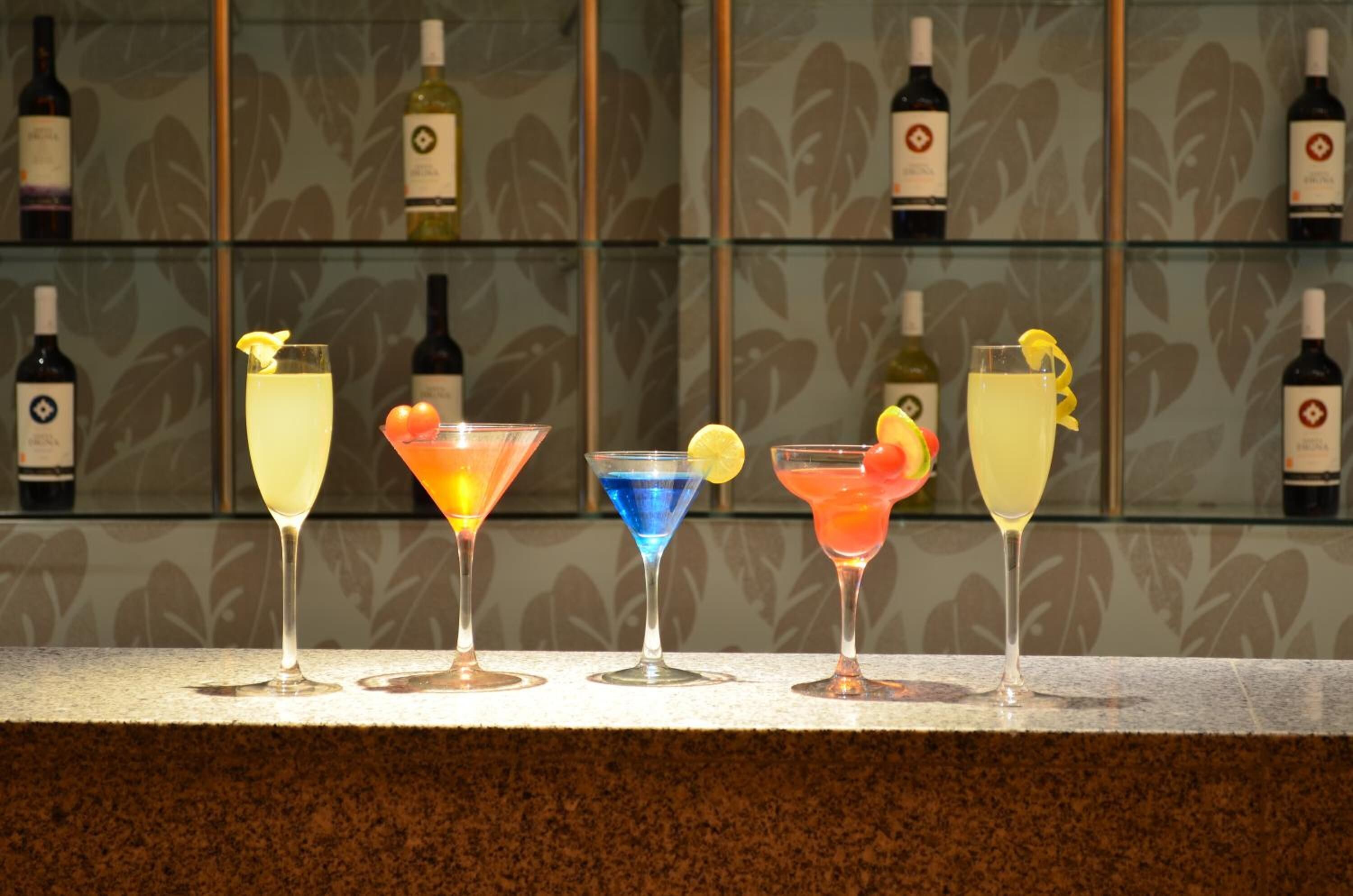Fem cocktails på en bardisk med tapetbaggrund og vinflasker på hylder.