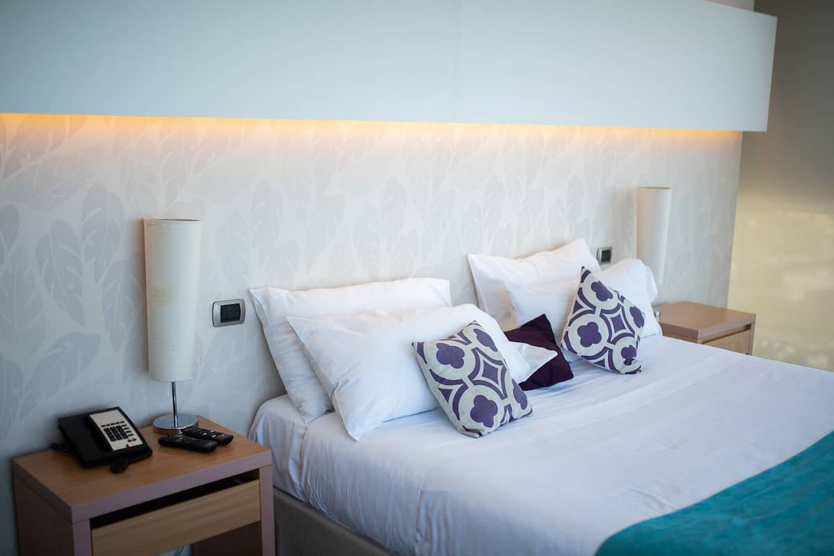 Un lit bien fait, avec du linge de lit blanc et des coussins à motifs, une lampe de chevet, un téléphone et une télécommande sur une table de chevet en bois.