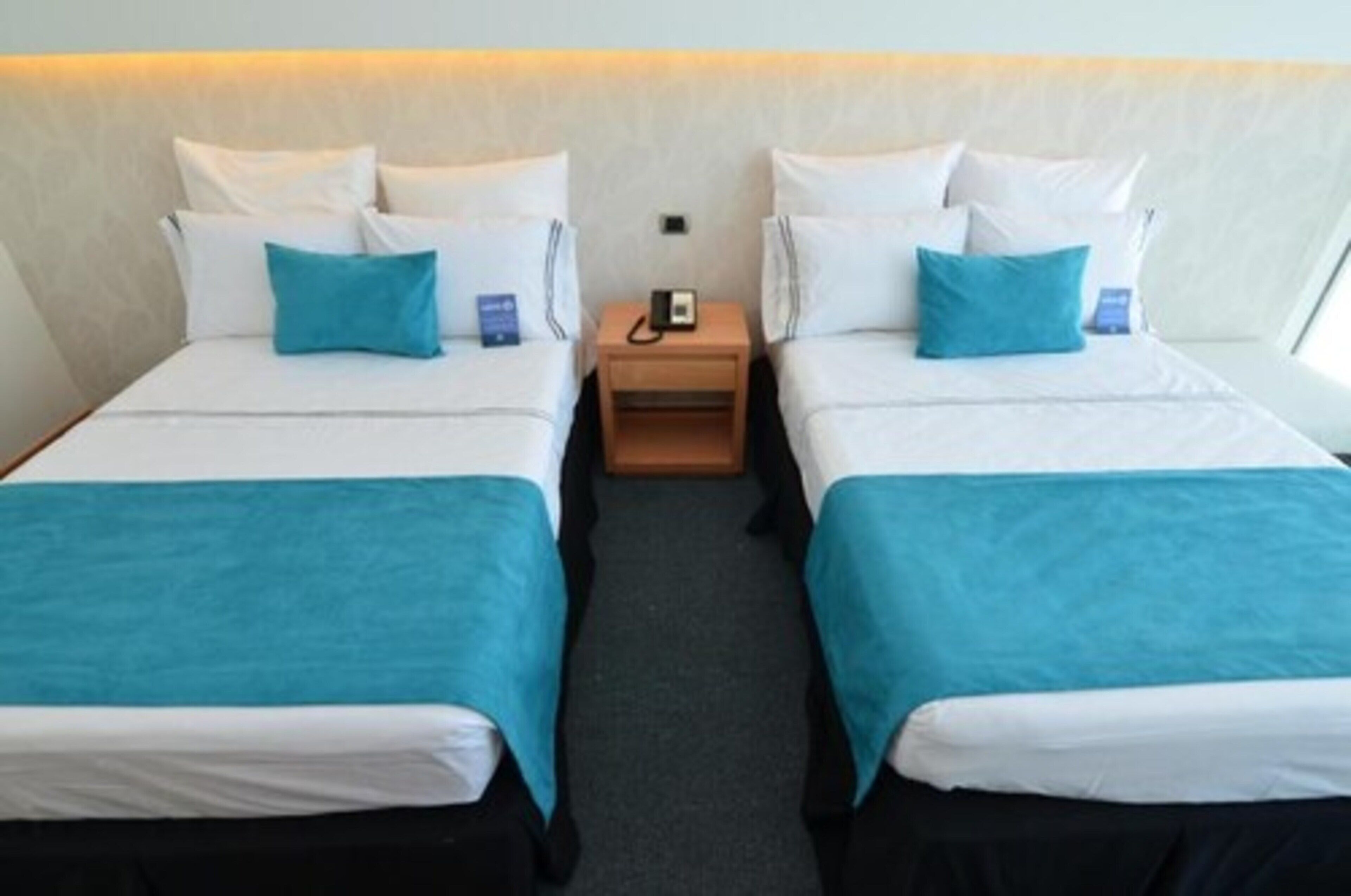 A hotel room with two beds, each with white bedding and teal throw pillows. There is a wooden nightstand between the beds with a phone and a small blue item on it.