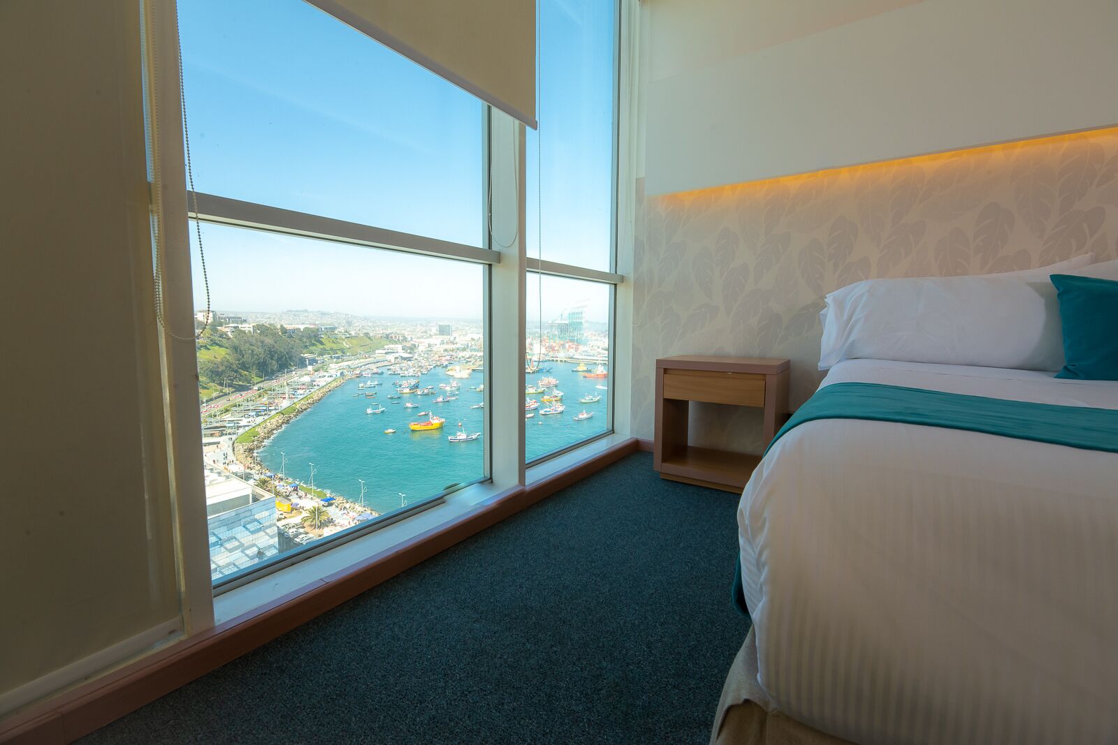 Quarto de hotel com uma janela ampla que oferece vista para um porto com barcos e o horizonte urbano.