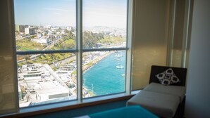 Quarto com uma janela grande que dá para a paisagem costeira, uma cama com travesseiro estampado e vista para barcos na água.