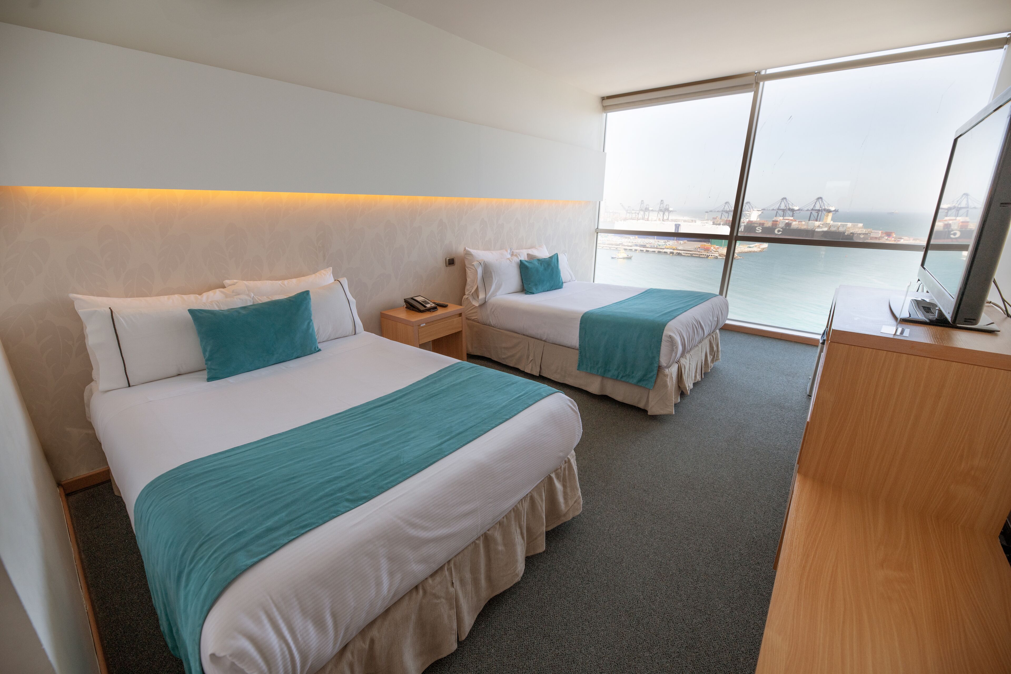 A hotel room with two beds, a desk, and a large window offering a view of the water and port.