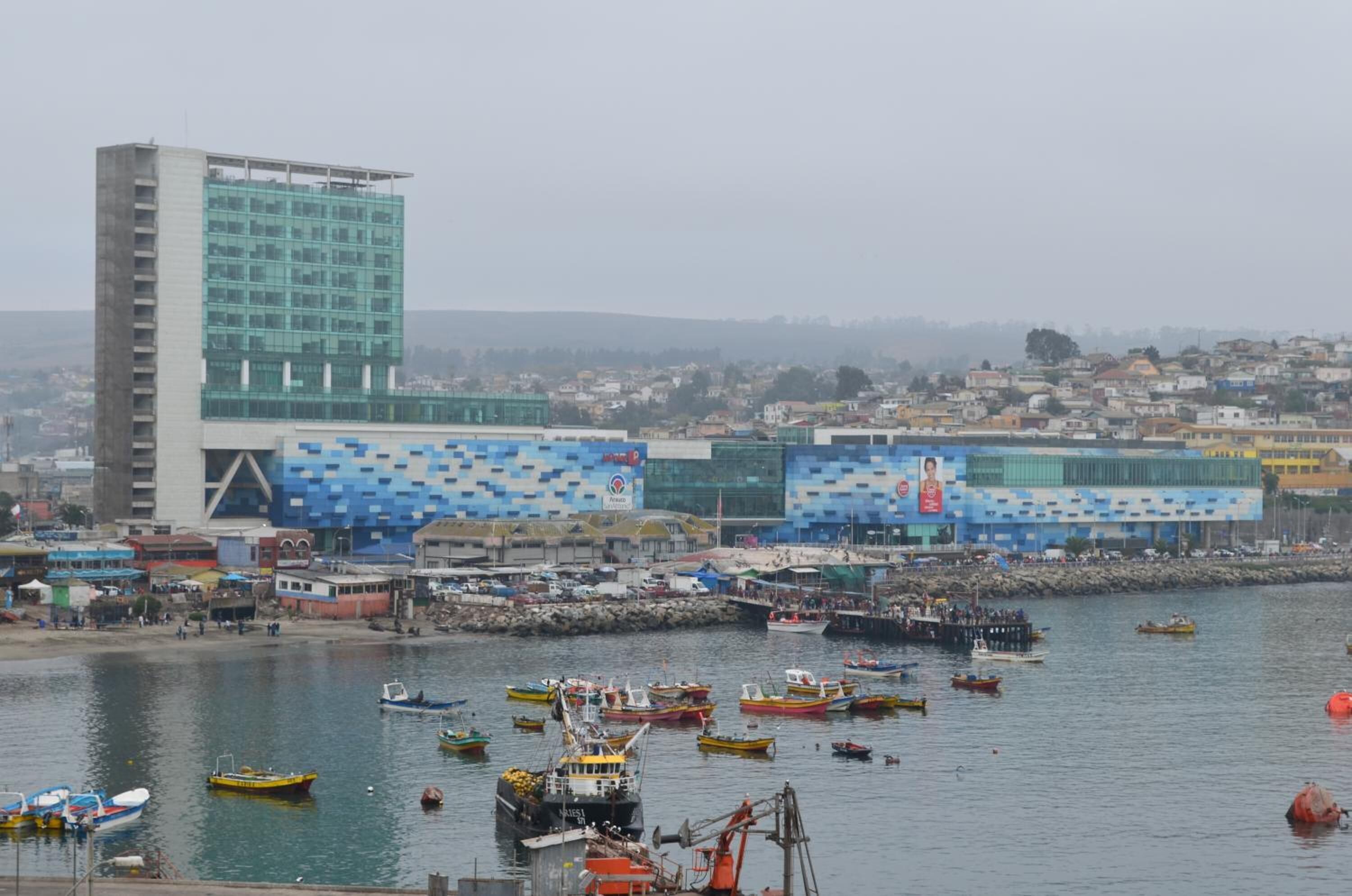 A coastal cityscape with a prominent modern building featuring a blue and white patterned facade, a marina with numerous boats, and a densely populated urban background.