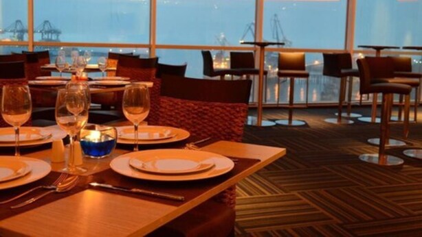 Um restaurante com mesas preparadas para refeições, grandes janelas e vista para a paisagem urbana.