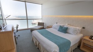 Quarto de hotel com uma cama grande, uma escrivaninha, uma cadeira e vista para o mar.