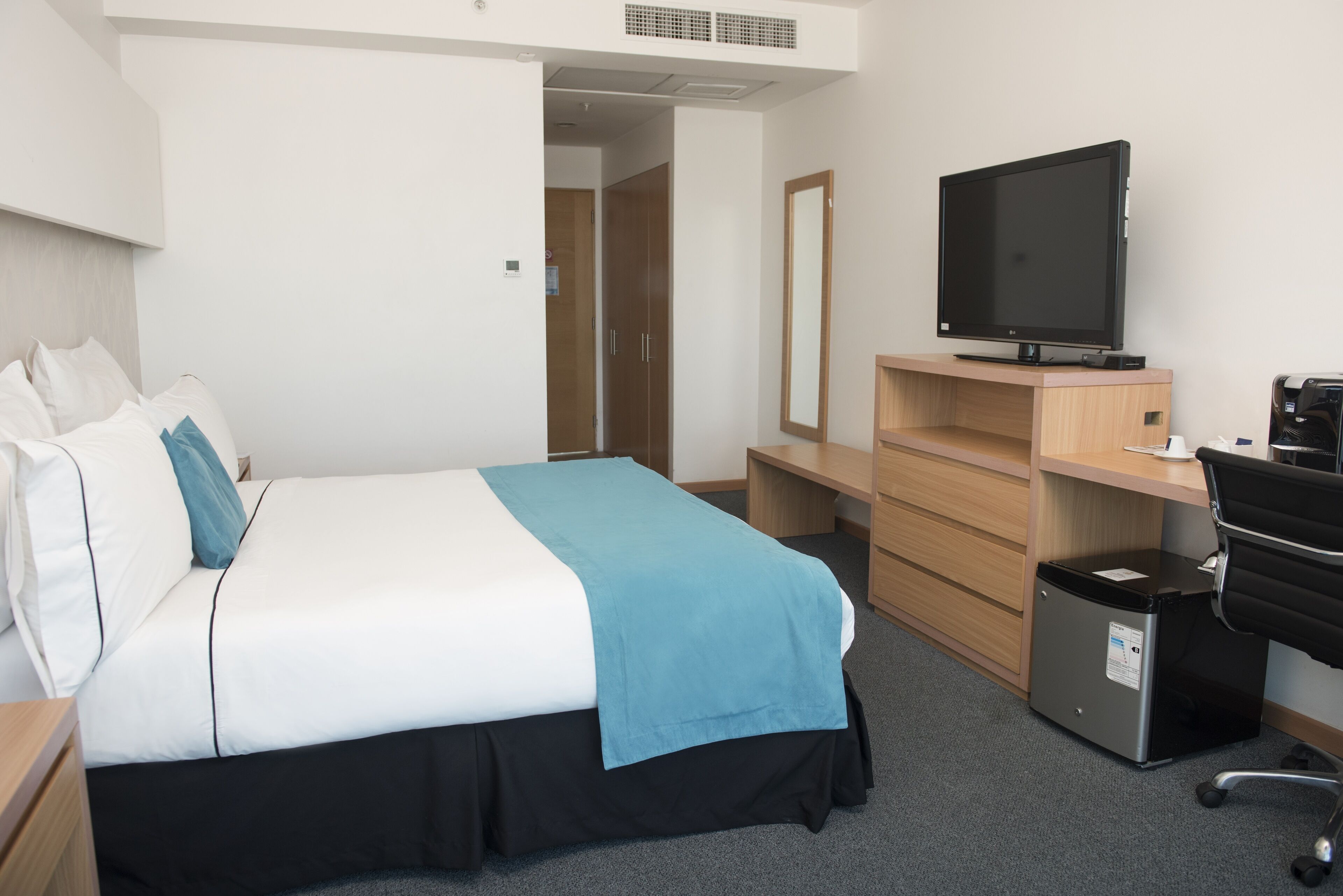 A hotel room with a bed, a desk, a TV, a mirror, and a trash bin.