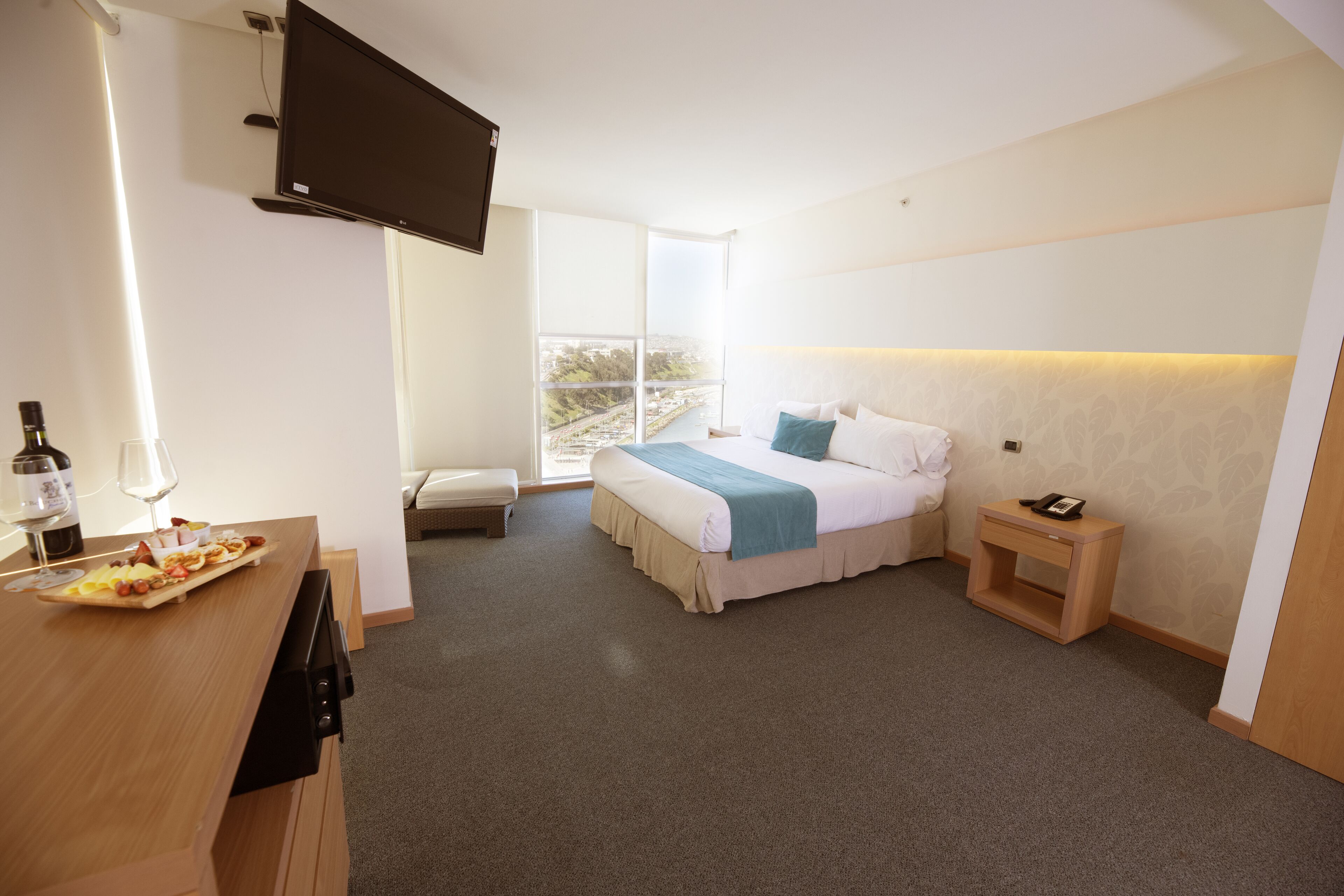 A hotel room with a bed, bedside table, a flat-screen TV mounted on the wall, a small window, and a breakfast bar with a plate of food.