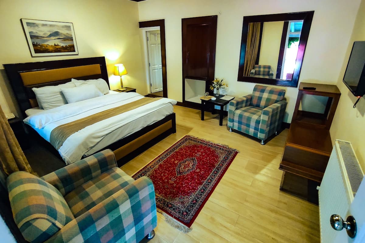 Une chambre d’hôtel avec un lit, deux fauteuils à motifs écossais, un tapis rouge, une petite table avec une lampe, un miroir et un téléviseur posé sur un meuble.