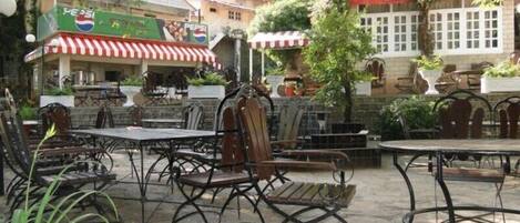 An outdoor seating area with metal tables and chairs, surrounded by greenery and a building with a red and white striped awning.