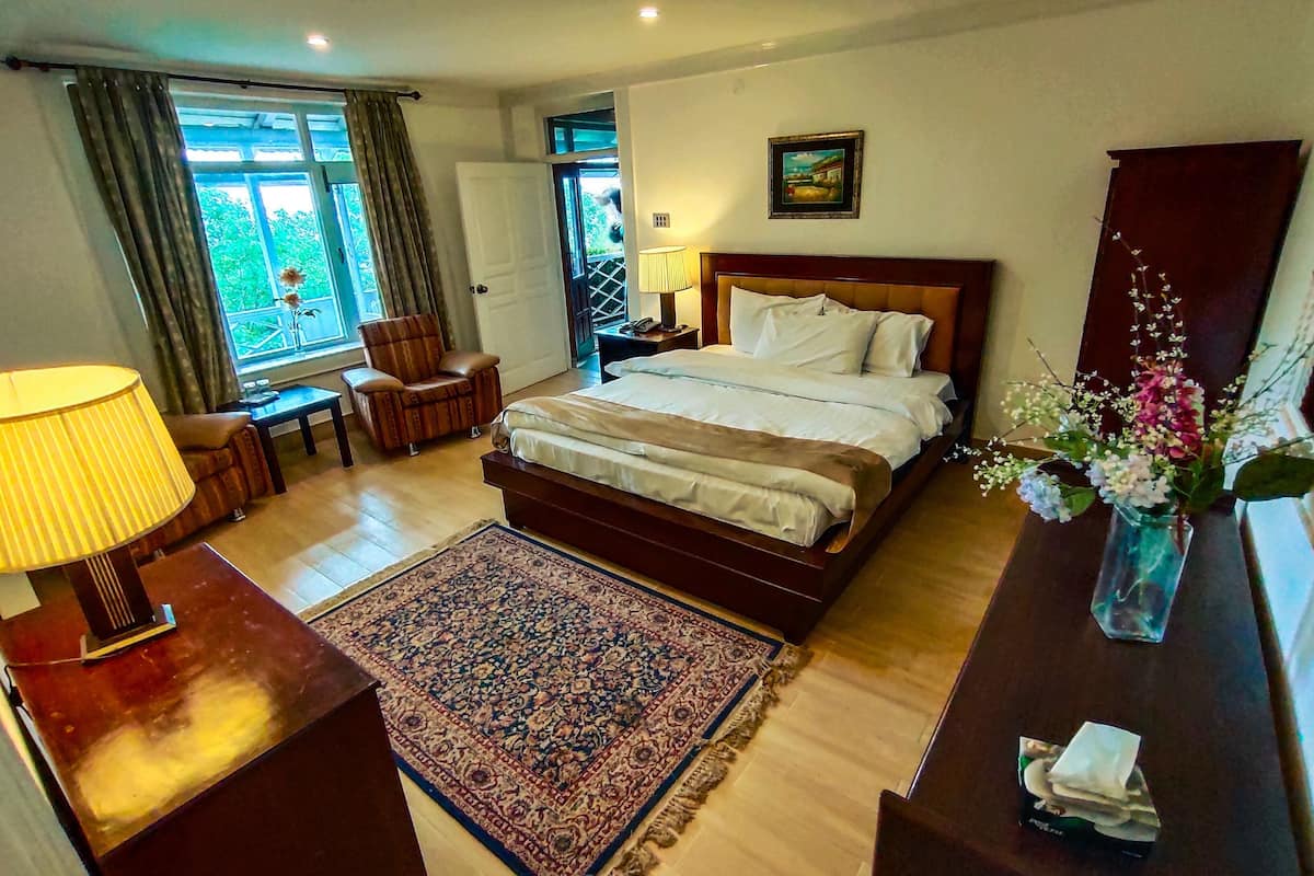 Une chambre d’hôtel avec un grand lit, un bureau avec une lampe, une chaise, un tapis et une fenêtre avec des rideaux.