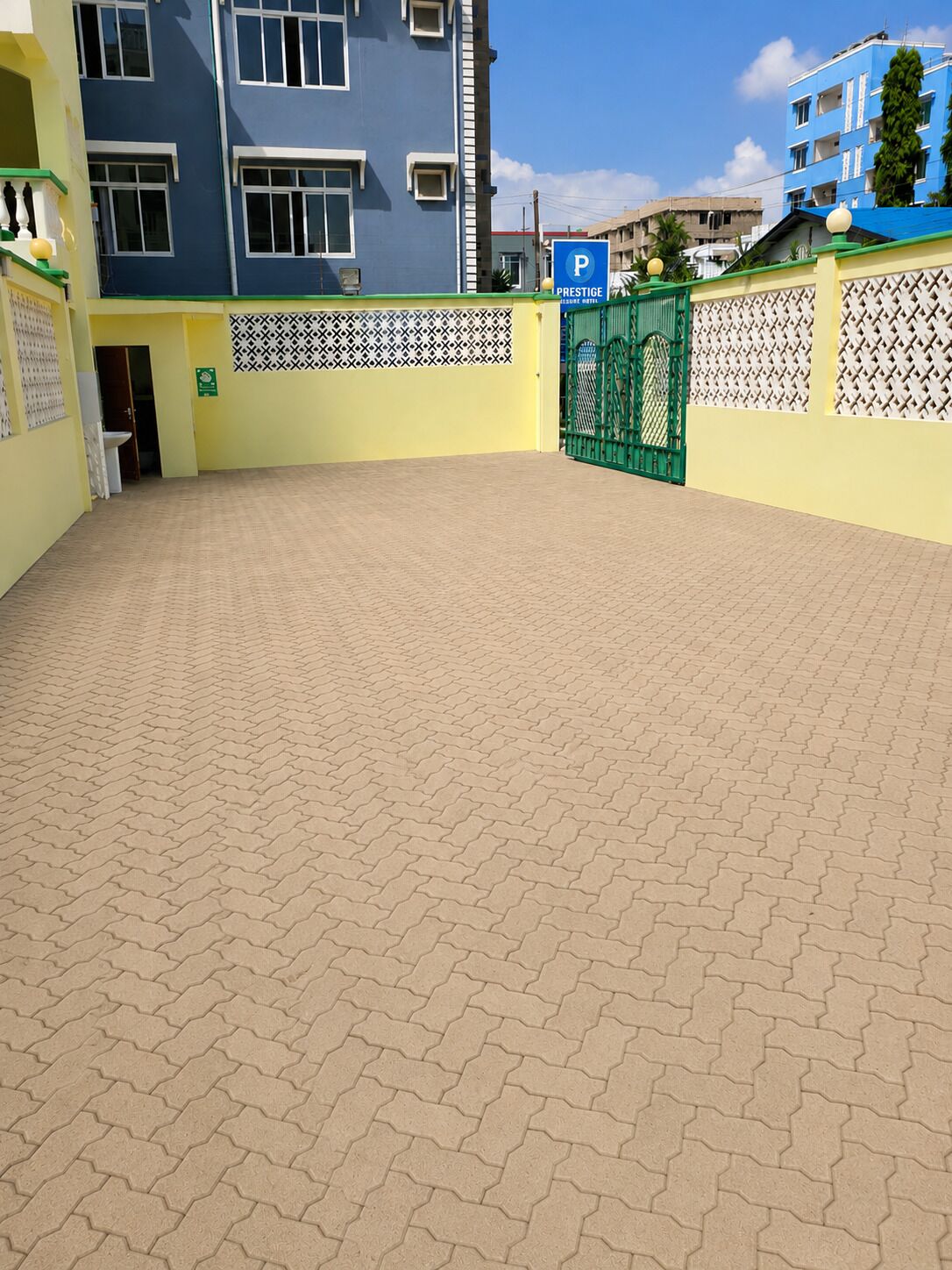 Um pátio com piso pavimentado, portão verde e um edifício azul com placa de estacionamento.