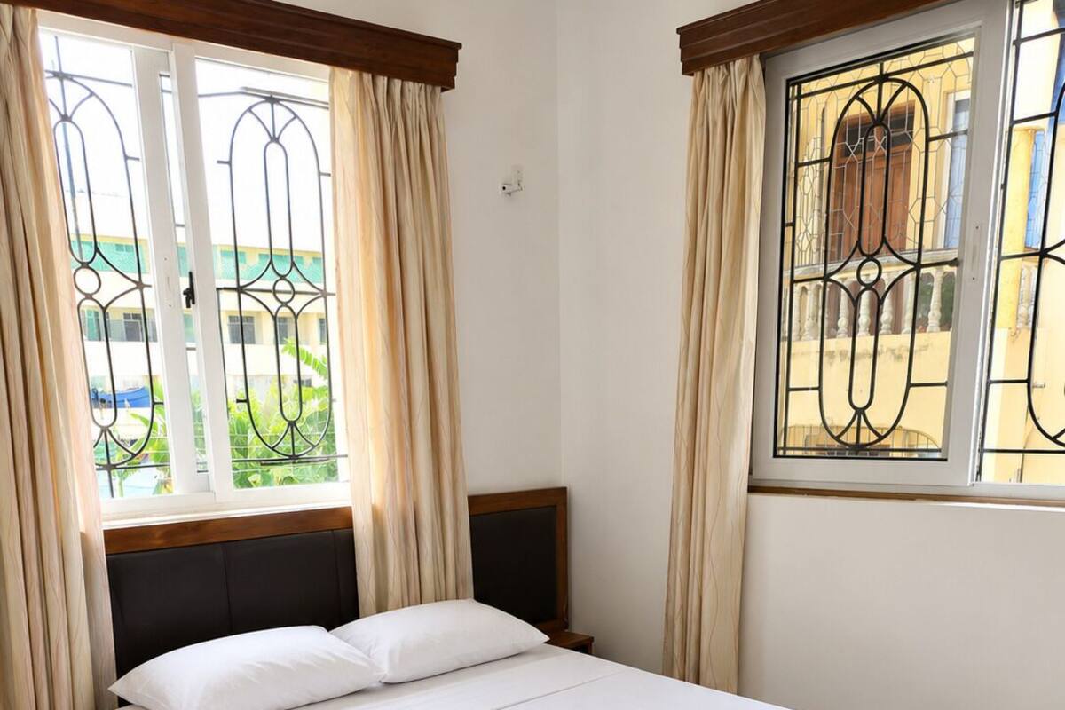 A single bed with a wooden frame and white bedding, two bedside tables, a window with curtains, and a view of greenery outside.