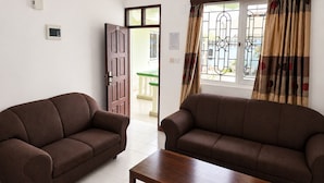 Une pièce avec deux canapés marron, une table basse en bois, un sol carrelé de couleur blanche, une fenêtre avec des rideaux et une porte donnant sur un balcon.