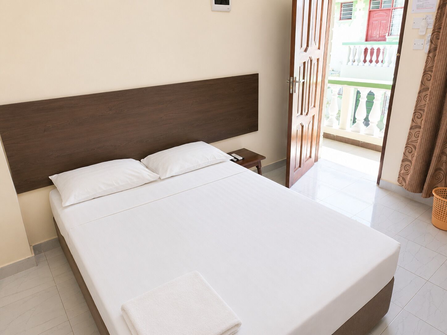 Un lit avec du linge de lit blanc, une tête de lit en bois, une table de chevet avec une télécommande, et une vue sur l’extérieur par une porte ouverte.