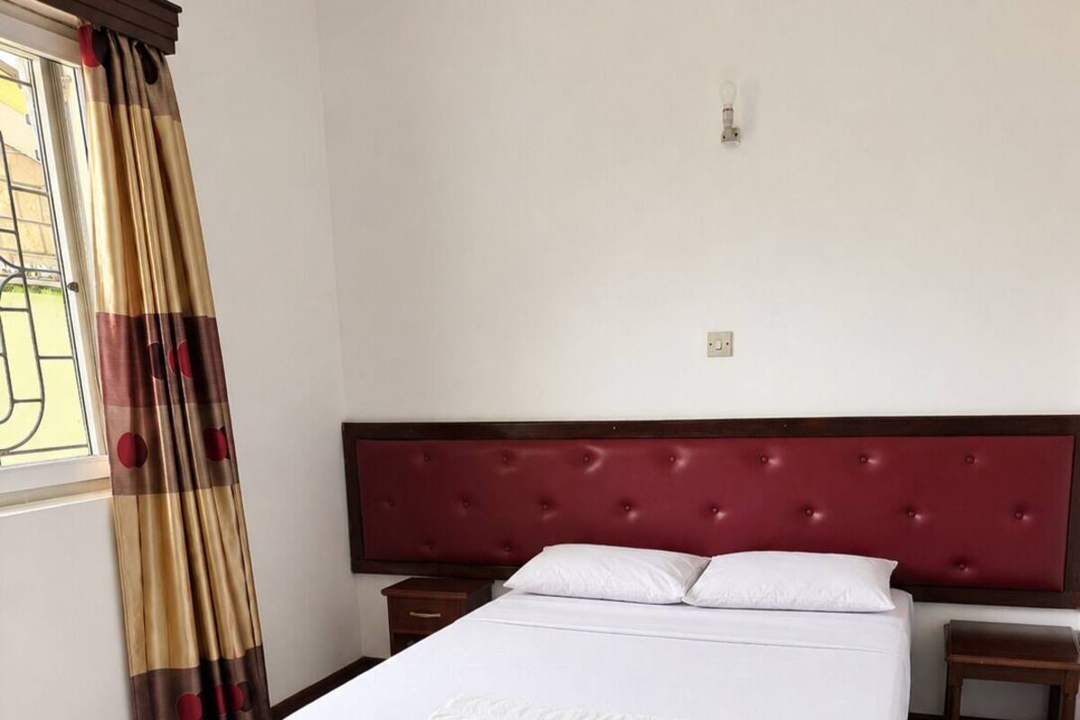 A hotel room with a large bed, a nightstand, a wooden headboard, and a window with curtains.