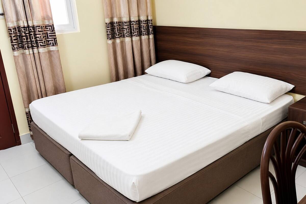 A neatly made bed with white linens, a wooden headboard, a bedside table with a remote control, and a chair.