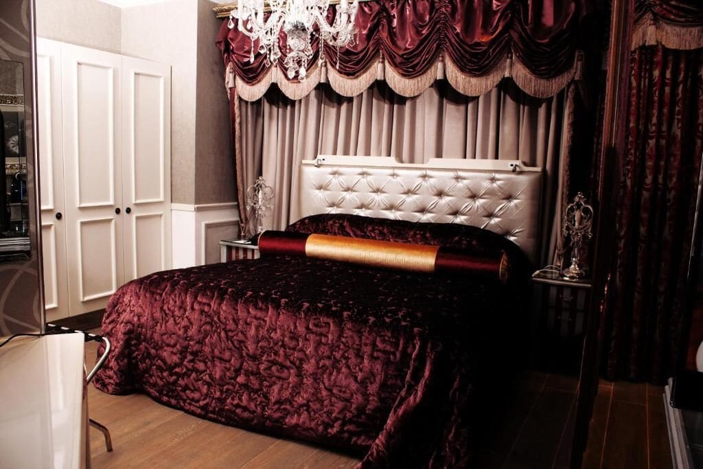 A bedroom with a tufted headboard, a large bed with a red blanket, a white wardrobe, and a chandelier.
