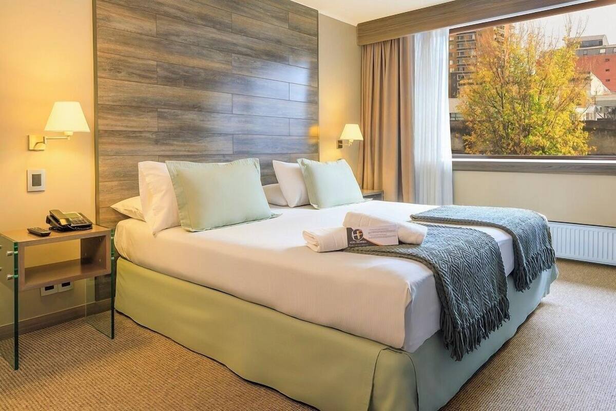 A hotel room with a bed, bedside lamps, a telephone, a nightstand, and a window with a view of buildings and trees.