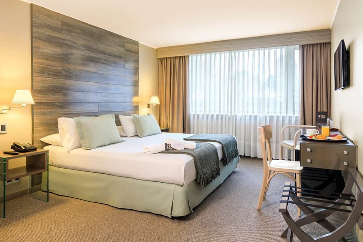 A hotel room with a bed, bedside tables, a desk with a chair, a TV, and a window with curtains.