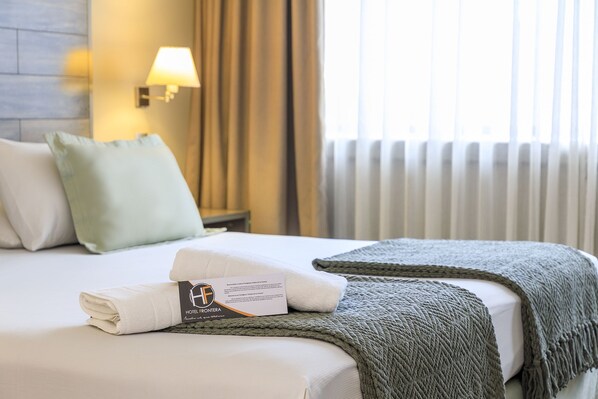 Una cama bien hecha con una toalla doblada y una colcha, una lámpara en la pared y una ventana con cortinas.