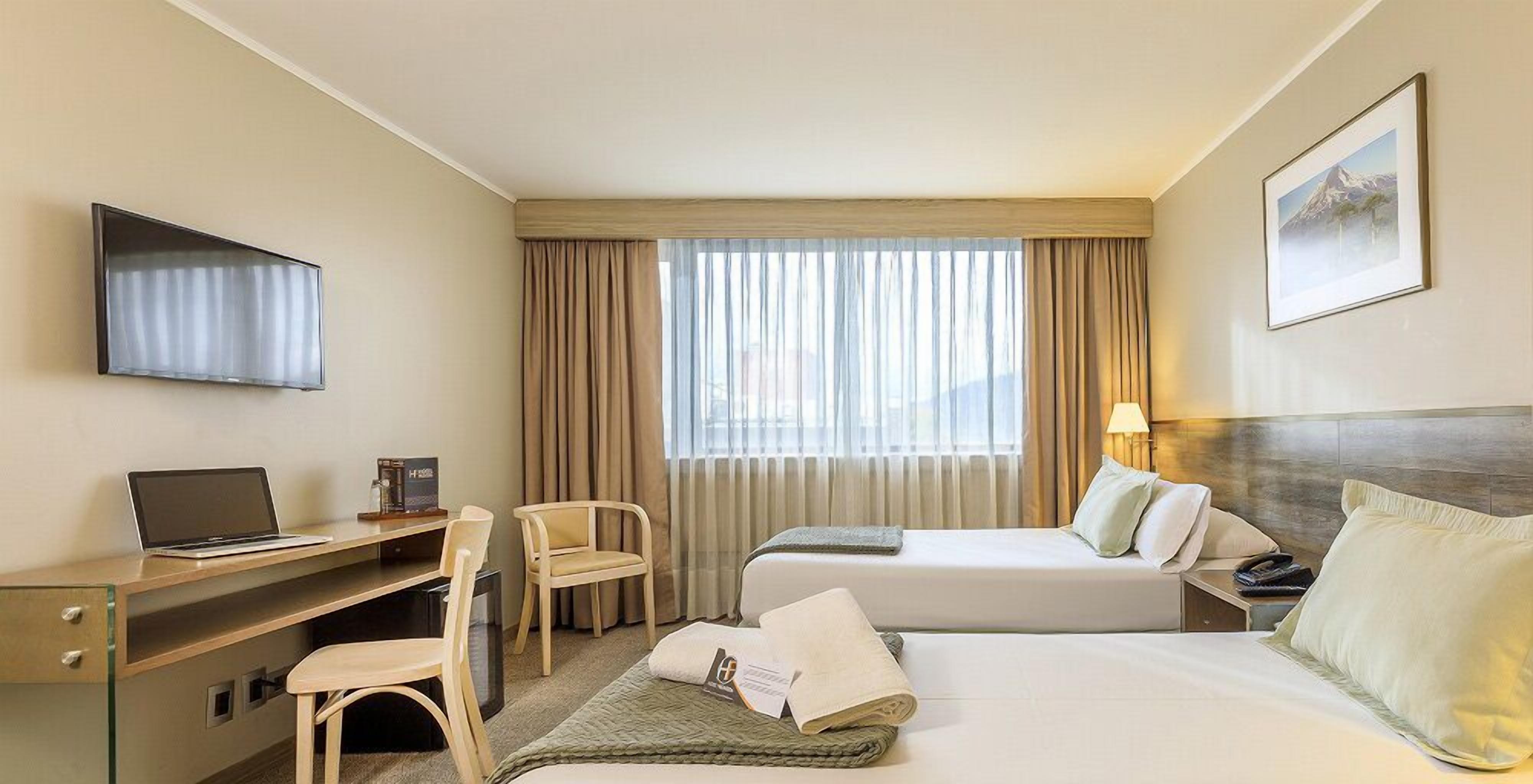 A hotel room with a bed, a desk with a laptop, a chair, a TV mounted on the wall, and a window with curtains.