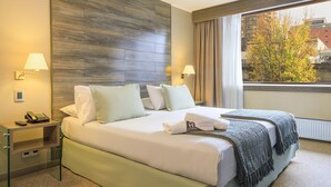 A hotel room with a large bed, two bedside lamps, a telephone, and a view of buildings and trees through the window. - Hotel Frontera Plaza (Temuco)