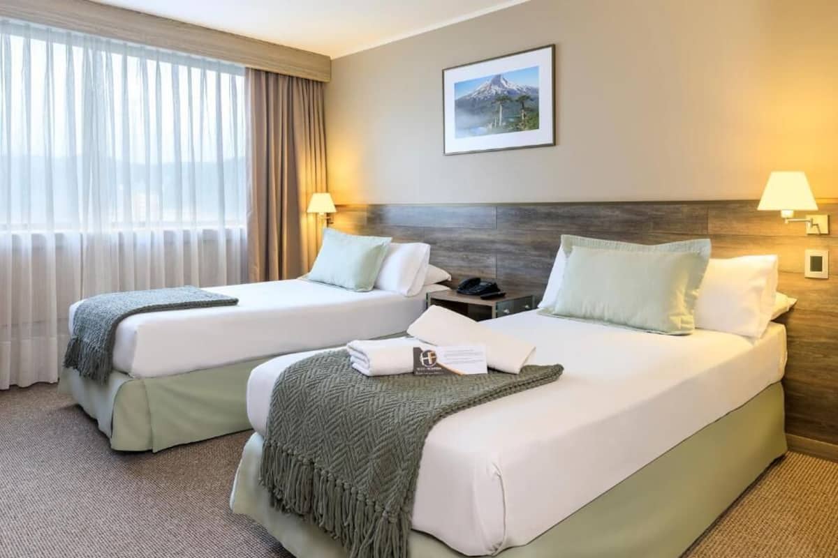 A hotel room with two beds, a large window with curtains, a framed picture on the wall, and two bedside lamps.