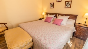 A neatly made bed with floral bedding and pink pillows, a wooden nightstand with a lamp, and a wooden bench. - Posada Real Quinta San José (Piedralaves)