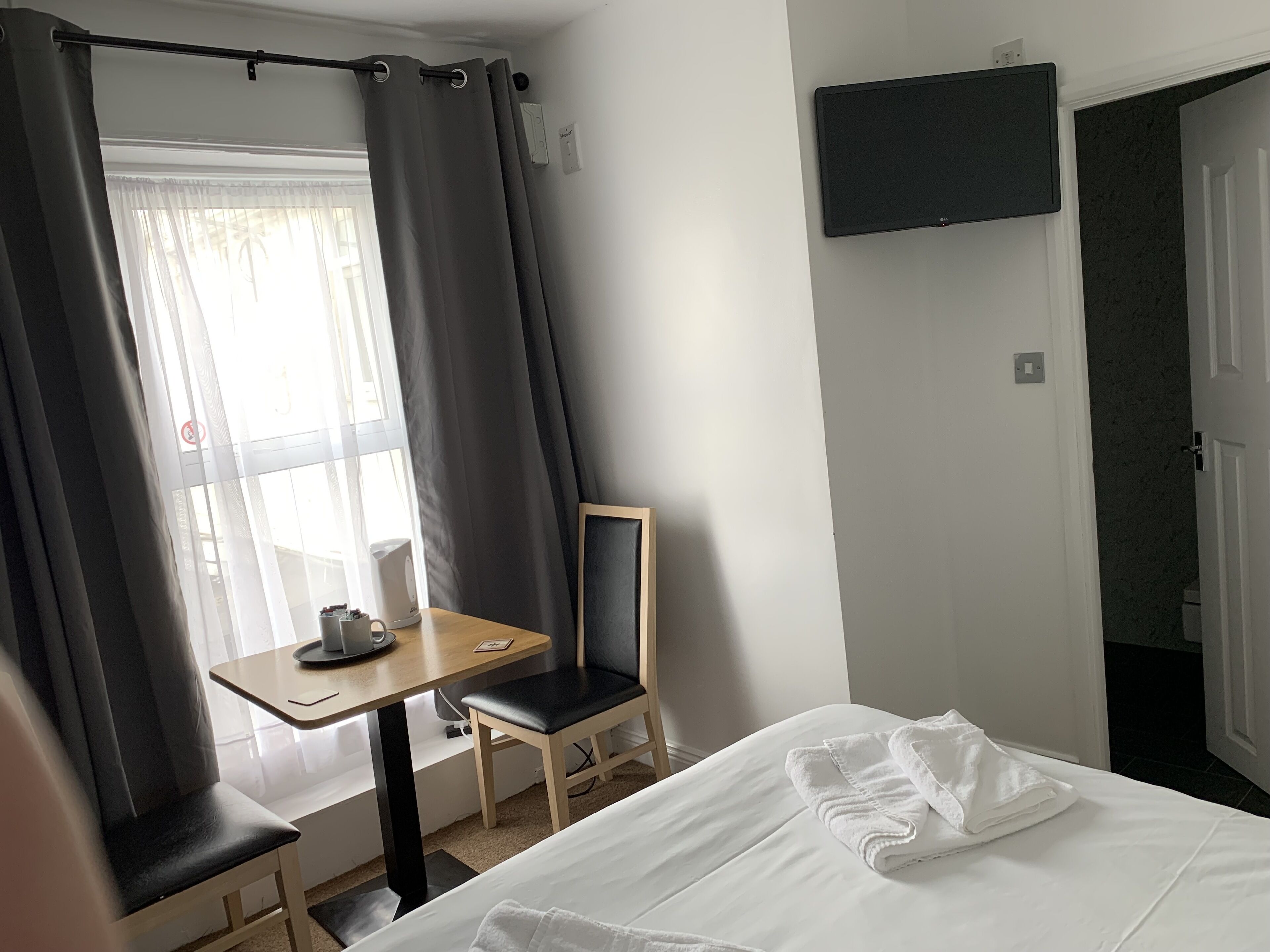 A hotel room with a bed, a desk with a chair, a television mounted on the wall, and a window with curtains.