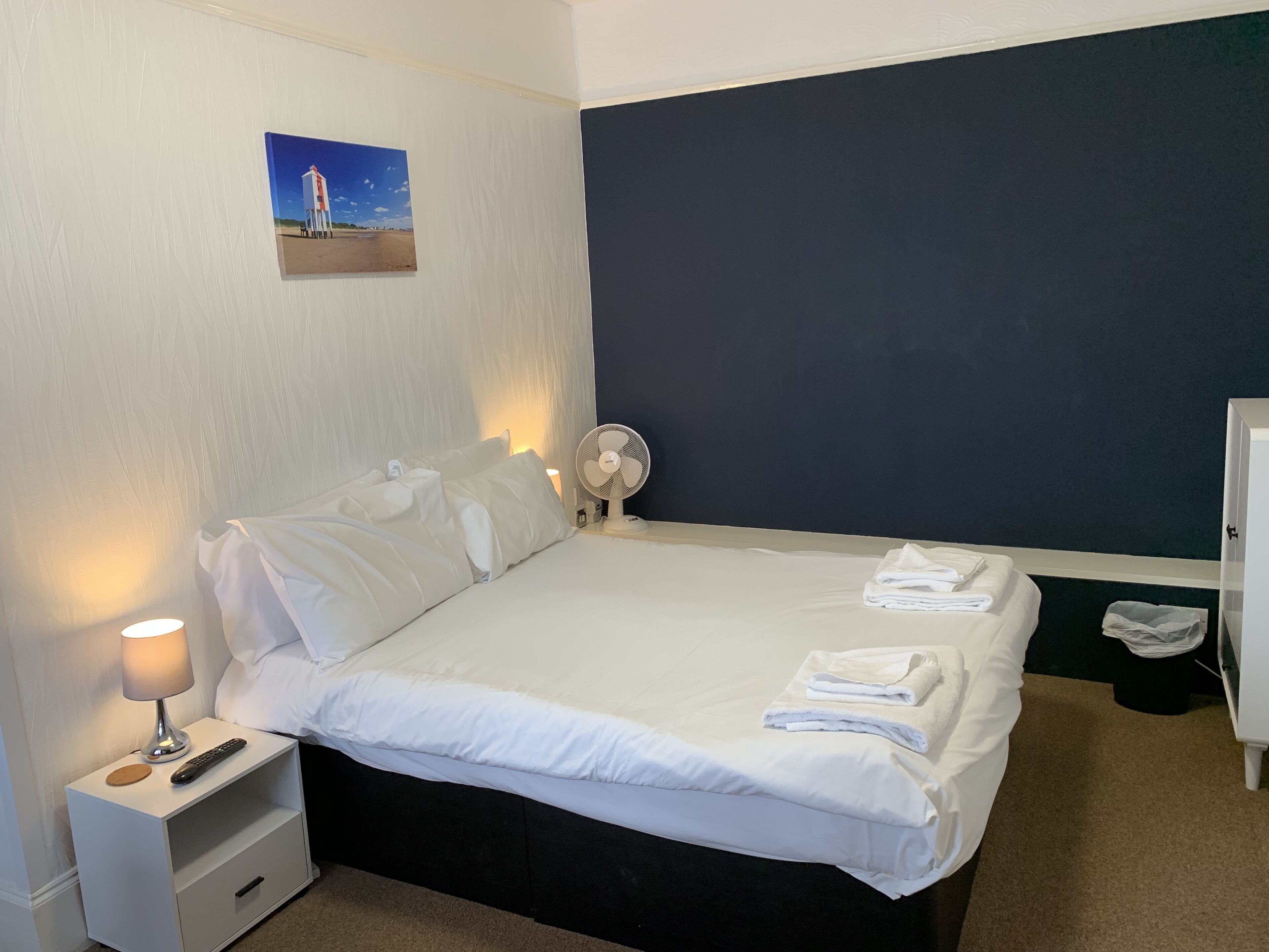 A neatly made bed with white linens, a bedside lamp, a remote control, and a folded towel on the nightstand.