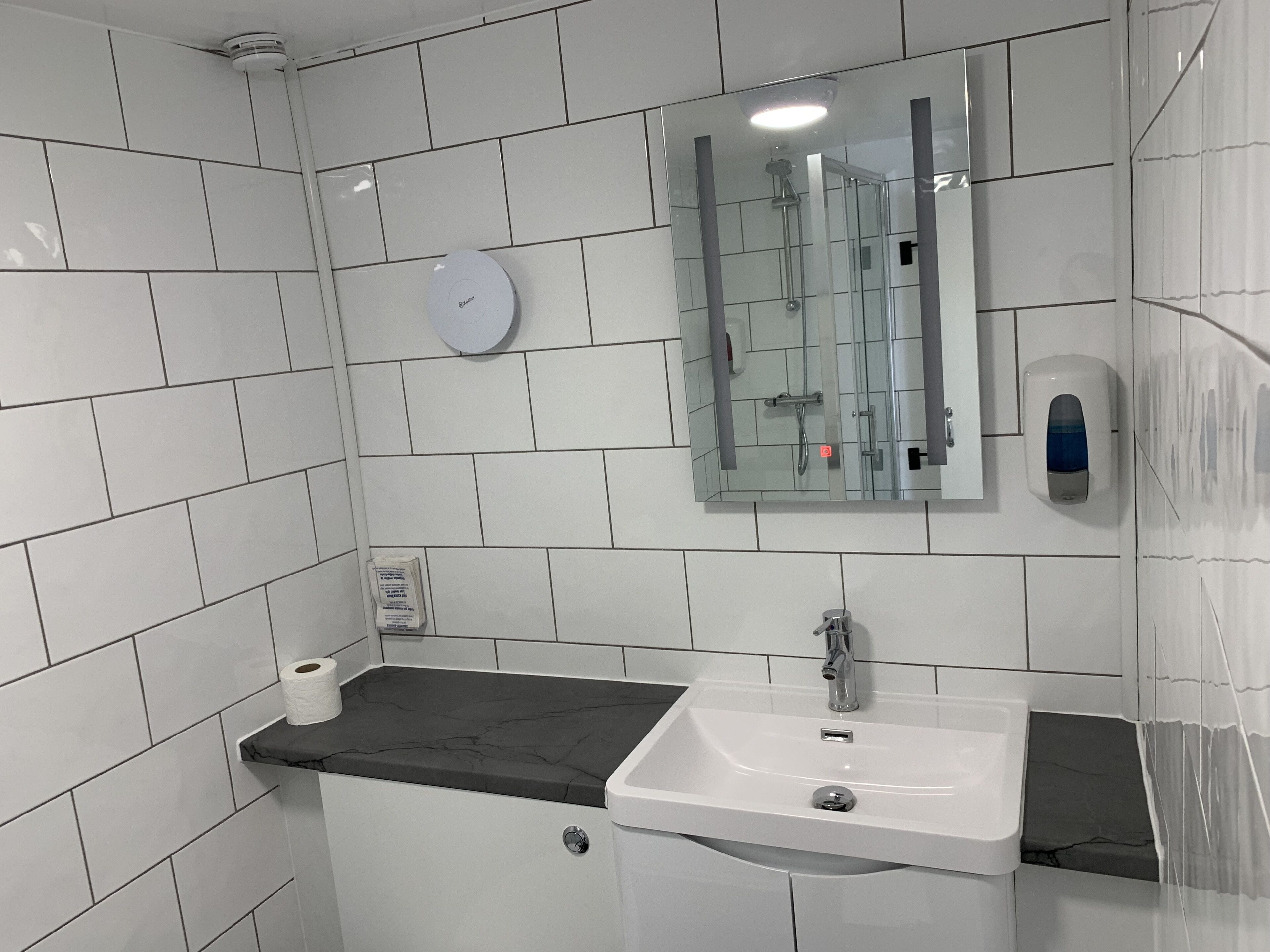 Uma casa de banho com paredes azulejadas em branco, uma pia branca com bancada preta, um espelho, um dispensador de sabonete para a mão e um dispensador de papel higiénico.