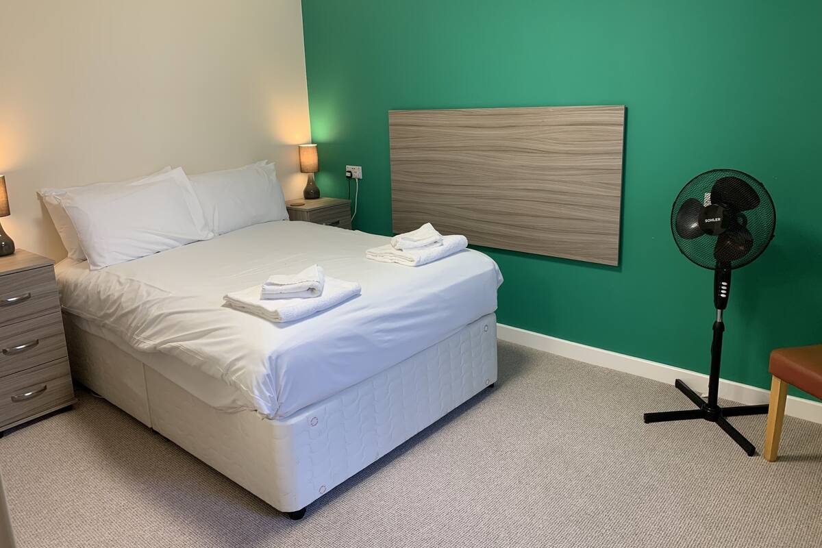 A neatly made bed with white linens and towels, a bedside lamp, a wall-mounted fan, and a wooden headboard.