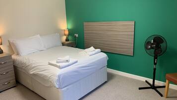 Uma cama bem arrumada com lençóis e toalhas brancos, uma luminária de cabeceira, um ventilador fixado na parede e uma cabeceira de madeira.