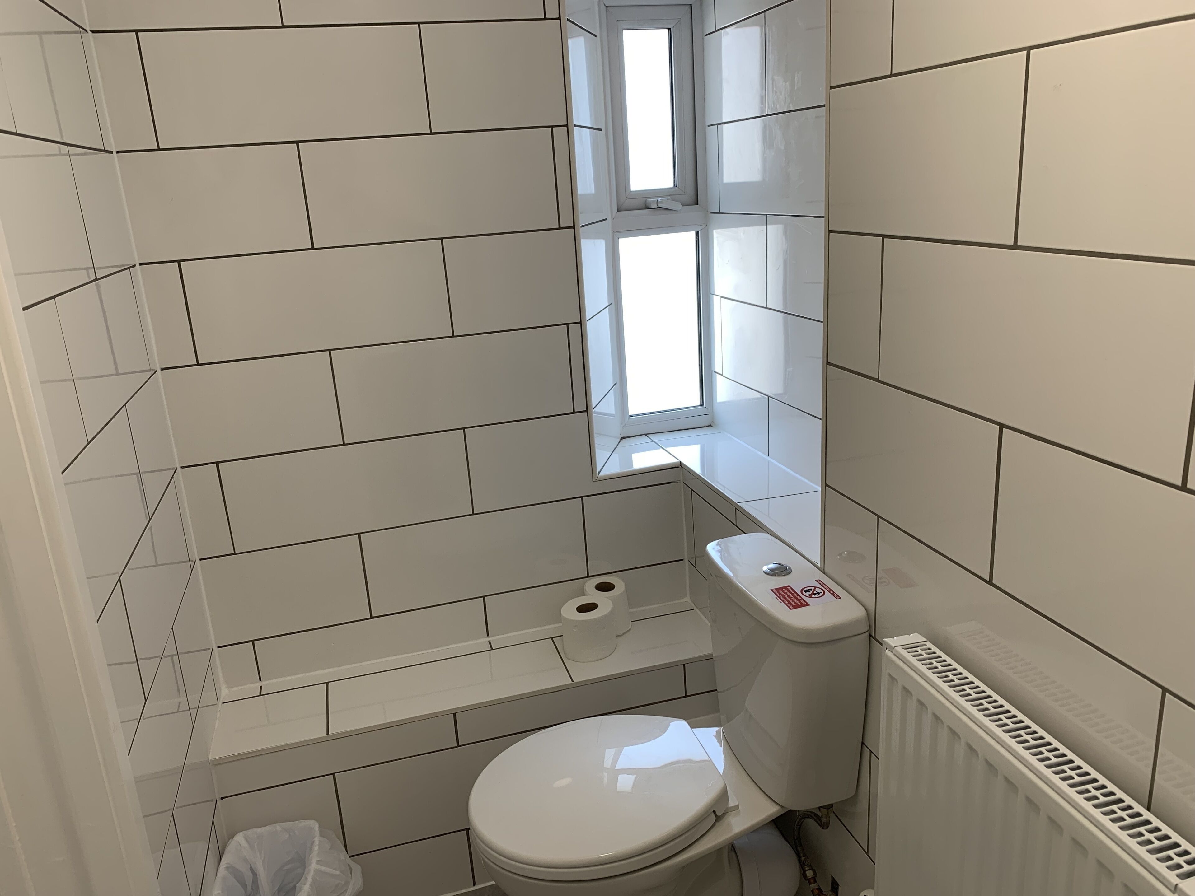 Une petite salle de bain aux dimensions réduites, avec des carreaux blancs, des toilettes, une fenêtre et un radiateur.